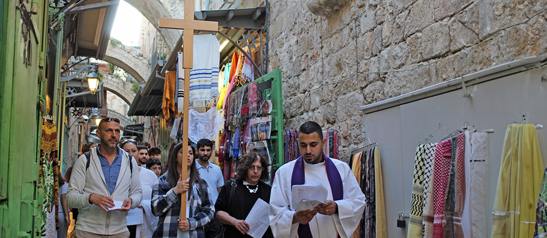 Mehrere Menschen auf einer Prozession durch die Altstadt Jerusalems. Im Vordergrund trägt eine junge Frau ein großes Holzkreuz.