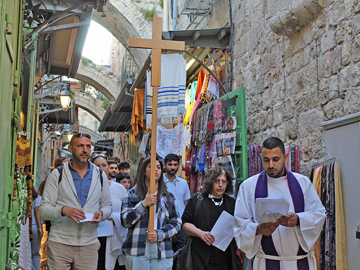 Mehrere Menschen auf einer Prozession durch die Altstadt Jerusalems. Im Vordergrund trägt eine junge Frau ein großes Holzkreuz.