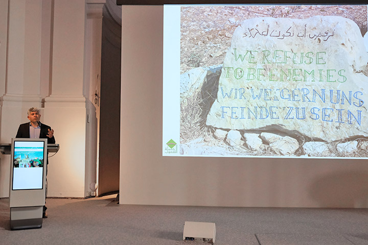Daoud Nassar steht an einem Pult und hält einen Vortrag. Auf einer großen Leinwand ist ein Foto mit einem dekorativ bemalten Stein. "Wir weigern uns Feinde zu sein", steht auf dem Stein.