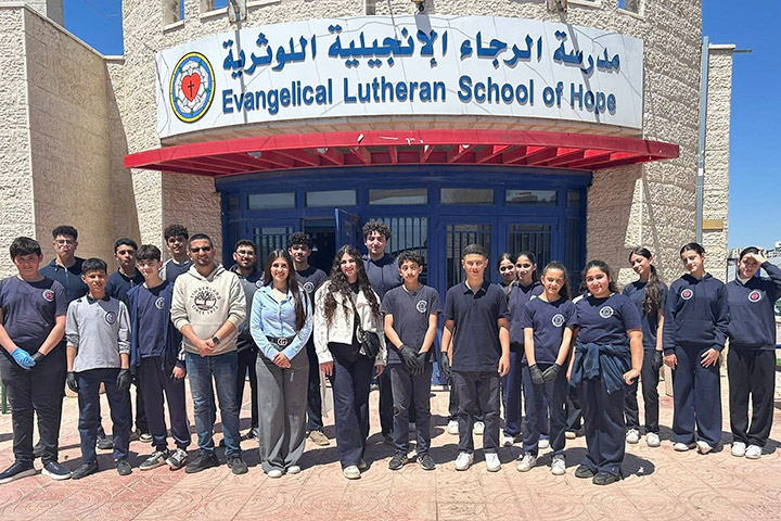 20 Schülerinnen und Schüler des Umweltclubs stehen vor dem Eingang der evangelischen Schule in Ramallah. Die meisten tragen dunkelblaue Shirts mit dem Logo der Schule. Über dem Eingang steht groß: Evangelical Lutheran School of Hope, auch in arabischen Schriftzeichen.