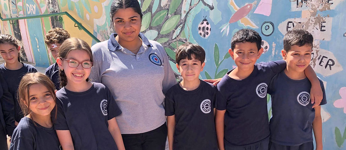 Auf dem Hof der evangelischen Schule in Ramallah stehen eine jugendliche Schülerin in einem grauen Shirt mit Schullogo und fünf deutlich kleinere Grundschüler mit blauen Shirts in einer Reihe. Einige legen den Arm auf die Schulter eines anderen.