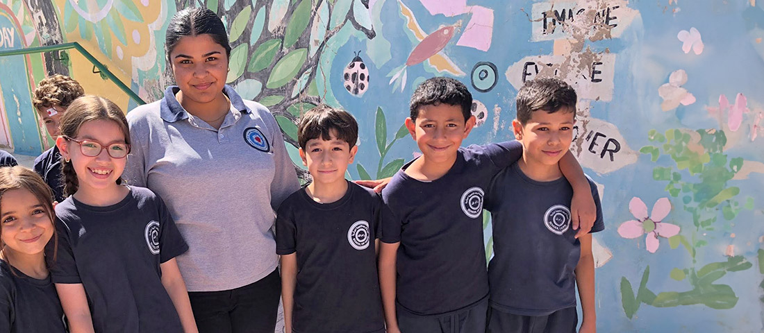 Auf dem Hof der evangelischen Schule in Ramallah stehen eine jugendliche Schülerin in einem grauen Shirt mit Schullogo und fünf deutlich kleinere Grundschüler mit blauen Shirts in einer Reihe. Einige legen den Arm auf die Schulter eines anderen.