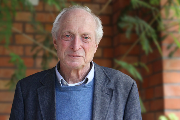 Jürgen Wehrmann, ein älterer Mann mit silbernen Haaren, bekleidet mit grauem Sakko, blauem Pullover und offenem Hemd, steht vor einer Wand aus Klinkersteinen.