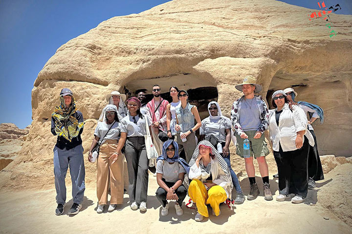 Eine Gruppe von etwa 15 jungen Erwachsenen mit heller und dunkler Hautfarbe, die meisten mit Kopfbedeckungen, vor einer Wüstenlandschaft  