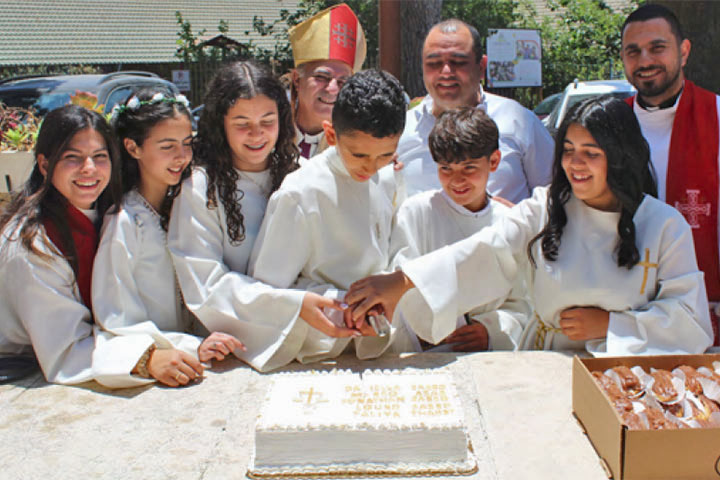 Die Konfirmanden, zwei Jungen und drei Mädchen in weißen Gewändern, schneiden eine Torte an. Im Hintergrund: Bischof Azar mit Bischofsmütze. Links und rechts stehen die jungen Geistlichen  Sally Azar und Rodney Said.