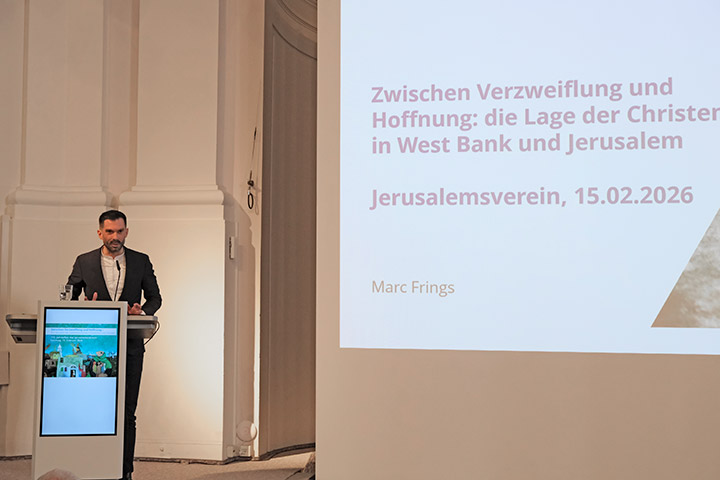Marc Frings steht links an einem Pult und hält seinen Vortrag. Links steht auf einer großen Leinwand das Thema der Veranstaltung.