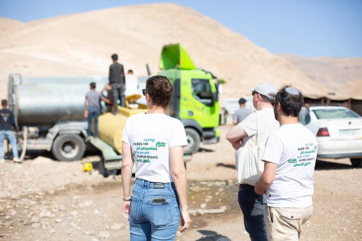 Drei Mitglieder der Rabbis for Human Rights stehen im Vordergrund, mit RHR-T-Shirts, mit dem Rücken zum Betrachter. Im Hintergrund ein Tankwagen und eine Wüstenlandschaft.