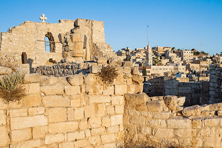 Hinter der Kirchenruine St. Georg mit einer verfallenen Mauer, auf der ein Kreuz steht, sieht man das Zentrum von Taybeh mit einem Kirchturm.