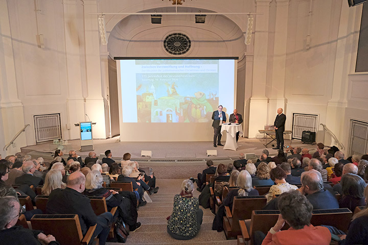 Auf dem Jahresfest des Jerusalemsverein sitzen etwa 200 Menschen in einem historischen Hörsaal. Auf der Bühne stehen Bischof Haddad, der Vorsitzende Wolfgang Schmidt und Matthias Wolf.
